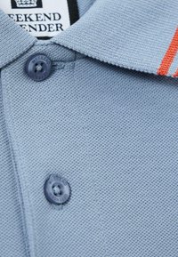 Svetlomodrá pletená polo košeľa s dvoma tmavomodrými gombíkmi označenými "Weekend Offender" a s oranžovou líniou na golieri.