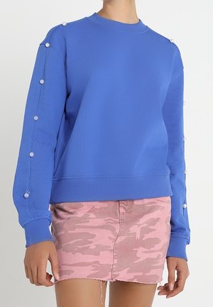 Blue long-sleeve sweater with pearl buttons along sleeves paired with a pink camouflage mini skirt on a standing person.