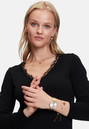 Zwarte trui met een V-hals en kantafwerking, gecombineerd met gouden sieraden, waaronder een horloge en gelaagde armbanden. Lichte achtergrond.