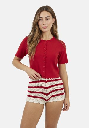 Vrouw met lang haar die een rood, kort mouwig, knoopsluitend shirt draagt en crèmekleurige gehaakte shorts met rode strepen, tegen een witte achtergrond.