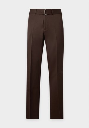 Uomo in piedi che indossa una giacca marrone sopra una camicia bianca, pantaloni marrone scuro e scarpe marroni su uno sfondo chiaro uniforme.