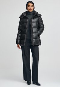 Svart, vadderad dunjacka med huva; har dragkedja och texturerat tyg. Bärs över en mörk polotröja och matchas med svarta byxor.