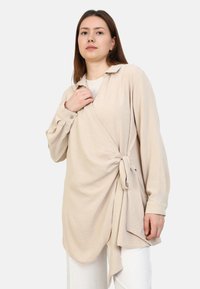 Beige wrap-paita pitkähihaisilla, eteen solmittavalla yksityiskohdalla ja kauluksella. Pehmeä kangas, jossa on sileä rakenne, ulottuu reiden puoliväliin.
