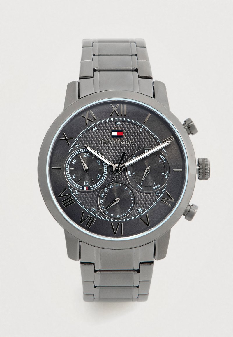 Orologio da polso Tommy Hilfiger con cinturino in metallo grigio, quadrante nero, numeri Romani, tre sottodials che mostrano funzionalità di 24 ore, giorno e data.