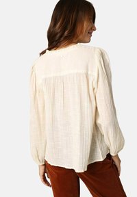 Crème-kleurige blouse van gestructureerde stof, lange pofmouwen, geplooide achterkant en een klein knoopdetail bij de hals. Gecombineerd met een paarse broek.