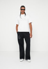 Λευκό κοντομάνικο μπλουζάκι με γραφικά κείμενα, συνδυασμένο με μαύρο παντελόνι φόρμ�ας με λογότυπο φλόγας και χαλαρή εφαρμογή. Λευκά αθλητικά παπούτσια.