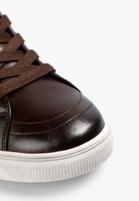 Zapatilla de cuero marrón con punta redondeada, superficie texturada y suela de goma blanca en contraste. Presenta cordones y costuras en marrón oscuro.
