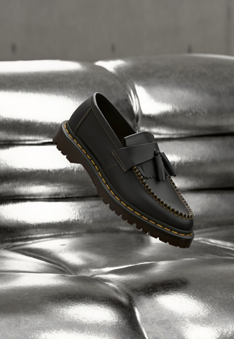 Μαύρο δερμάτινο μοκασίνι με φούντες και κίτρινα ράμματα που αιωρείται πάνω από ένα λαμπερό ασημί δερμάτινο καναπέ με κουμπιά.