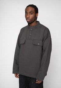 Ciemnoszary pulower z wysokim kołnierzem, z pięcioma guzikami i dwoma przednimi kieszeniami. Wykonany z miękkiego, teksturowanego materiału o luźnym kroju.