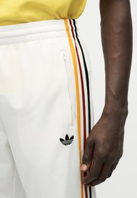 Κοντινό πλάνο λευκών παντελονιών φόρμας με πλευρικές ρίγες σε κίτρινο, κόκκινο και μαύρο χρώμα, τσέπη με φερμουάρ, λογότυπο Adidas, φορεμένα με κίτρινο πουκάμισο και εμφανές χέρι.