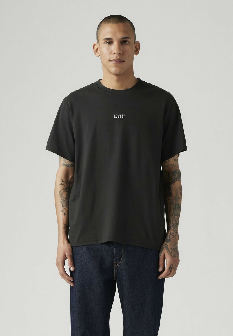 Joven con el cabello corto, vestido con una camiseta negra de Levi's y vaqueros azul oscuro, mostrando tatuajes en ambos brazos, de pie frente a un fondo liso.