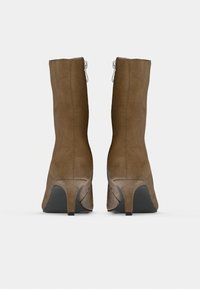 Bottines en daim marron avec une silhouette ajustée, un talon angulaire et un détail de fermeture éclair argentée. Texture lisse, légèrement resserrée en haut.