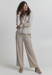 Beige gestreifte Bluse mit blauen Linien und lockerer Passform, kombiniert mit dazu passenden weiten Hosen und grünen Fransen-Blockabsatzsandalen.