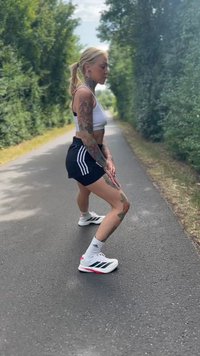 Urheilullinen tatuoitu nainen venyttelee jalkojaan asfaltilla, jota reunustavat vihreät puut, pukeutuneena urheiluvaatteisiin ja valkoisiin Adidas-kenkiin.