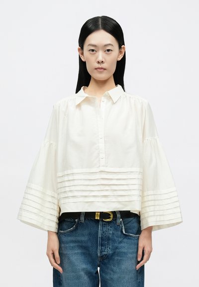 Kvinde med langt sort hår iført en løs hvid bluse med vandrette plisseringer og blå jeans med sort bælte mod en ensfarvet baggrund.