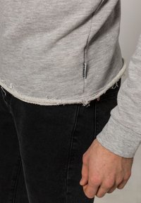 Chemise à manches longues gris clair avec des détails d'ourlet brut, portée sur un jean noir, avec une petite étiquette cousue sur le côté.