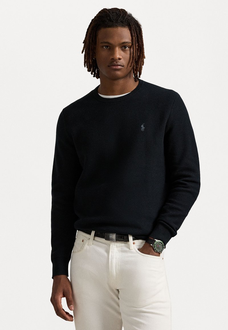 Sweatshirt en tricot noir avec un col rond et des poignets côtelés, associé à un jean en denim blanc et une montre avec un cadran vert.