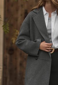 Manteau en laine gris avec larges revers, fermeture à boutons et manches larges, porté sur un pull gris et une chemise blanche boutonnée.