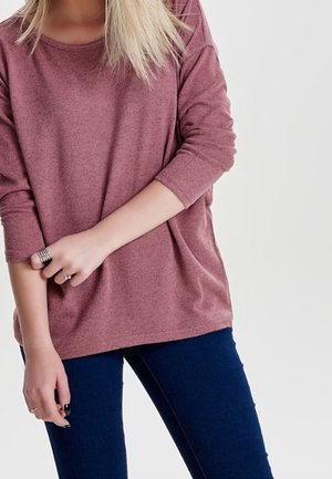 Femme aux cheveux blonds portant un haut mauve à manches longues et un pantalon bleu foncé, ajustant la manche gauche avec sa main droite.