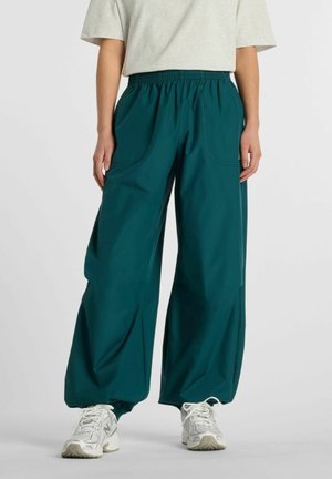 Personne portant un pantalon ample turquoise avec taille et poignets élastiques, associé à des baskets blanches et un haut à manches courtes gris clair.