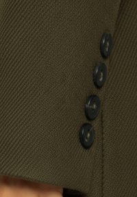 Närbild av olivgrön tyg med en texturerad yta och fyra svarta knappar som detalj längs kanten.