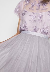 Laventelikjole med gennemsigtig, broderet liv, der har blomster mønstre. Tylleskørt prydet med små, hvide stjernelignende detaljer og satinbælte.