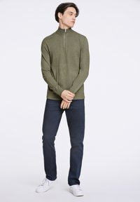 Suéter verde oliva de manga larga y cuello con cremallera, con un acabado texturizado, combinado con pantalones azul marino y zapatillas blancas.