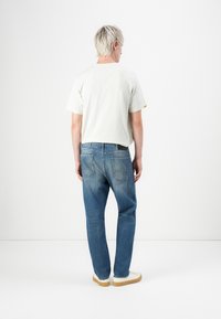 Hvid t-shirt med korte ærmer, blå denimjeans og hvide sneakers med en beige sål; synlige sømdetaljer på jeansene.