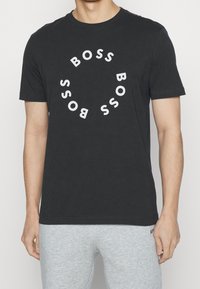 Man med svart t-shirt med vit "BOSS" text arrangerad i ett cirkulärt mönster och ljusgrå byxor mot en enfärgad bakgrund.
