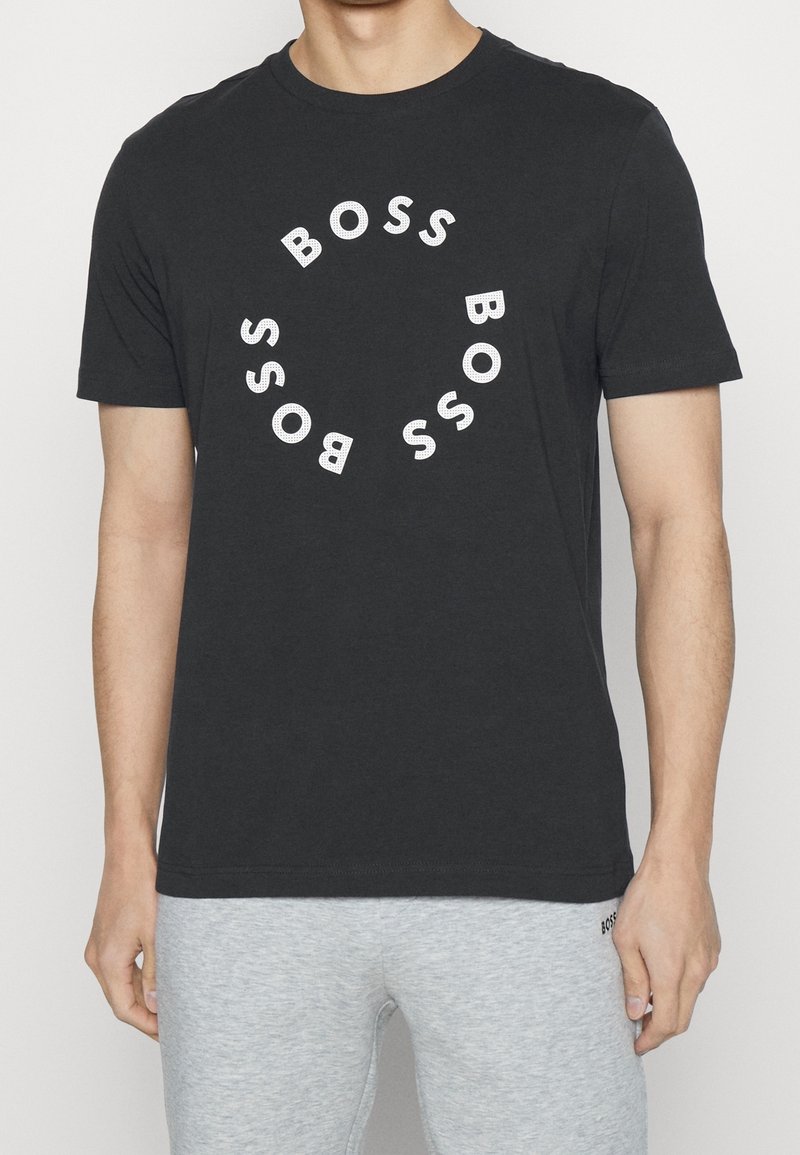 Man med svart t-shirt med vit "BOSS" text arrangerad i ett cirkulärt mönster och ljusgrå byxor mot en enfärgad bakgrund.