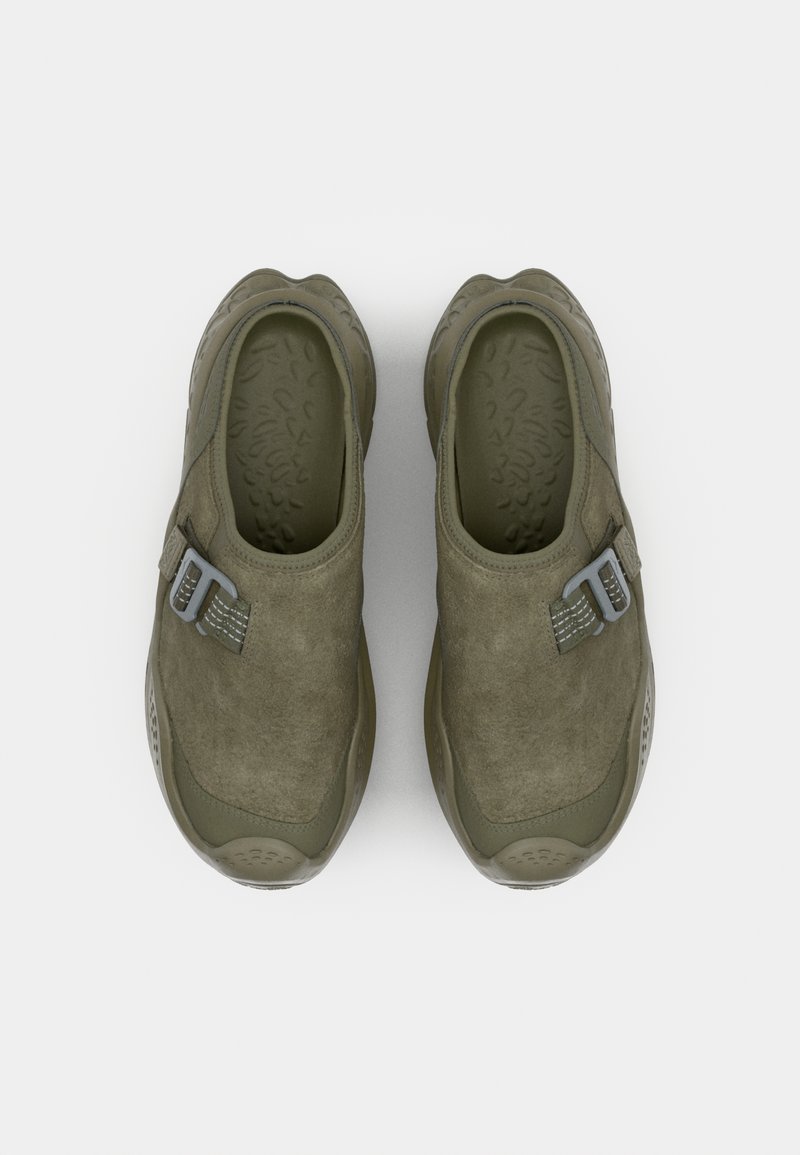 Olive green slip-on shoes with a smooth suede upper, rubber sole, and a light grey adjustable strap for fit. Textured insole detail visible.