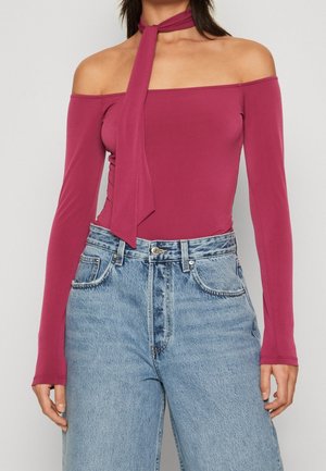 Femme portant un haut ajusté magenta à manches longues, épaules dénudées avec un nœud au cou, associé à un jean en denim bleu clair taille haute.