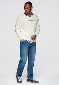 Hombre vestido con sudadera blanca con ribete verde, jeans azules y zapatillas navy, de pie con una mano en el bolsillo mirando de lado.