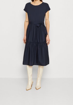 Femme portant une robe midi bleu marine avec une ceinture nouée à la taille et des manches courtes, associée à des bottes blanches montant jusqu'au genou, debout devant un fond uni.