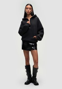 Czarna bluza z kapturem o luźnym kroju i przednią kieszenią, zestawiona z błyszcząca czarną mini spódnicą. Wysokie czarne botki z bocznymi guzikami dopełniają całość.