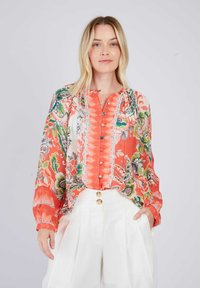 Bedruckte Bluse in Koralle und Weiß, mit floralen Mustern, Knöpfen an der Vorderseite und langen Ärmeln. Kombiniert mit hochgeschnittenen weißen Hosen.