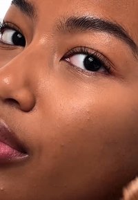 Nahaufnahme eines Gesichts mit glatter, gebräunter Haut, einem subtilen Schimmer, dunklen braunen Augen sowie gut definierten Wimpern und Augenbrauen.