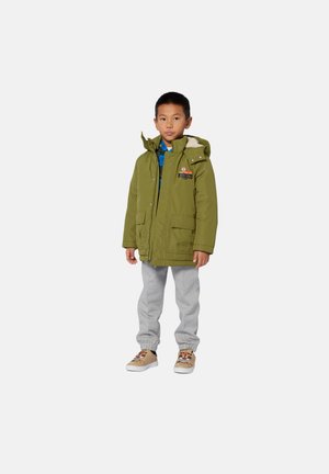 Un jeune garçon se tient debout, portant une veste à capuche verte, une chemise à motifs bleus, un pantalon de survêtement gris et des baskets beiges avec des visages de tigre.