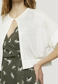 Cardigan bianco corto con maniche corte, dotato di un dettaglio a occhio di bue sul colletto; indossato sopra un vestito a stampa floreale verde.