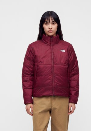 Giovane donna che indossa una giacca trapuntata bordeaux con collo alto e pantaloni beige, in piedi davanti a uno sfondo bianco uniforme.