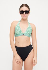 Zeleni kupaći top s halter izrezom i cvjetnim uzorkom, s logom Tommy Hilfigera na traci; uparen s visokim crnim kupaćim gaćicama. Nosi minimalističke sunčane naočale.