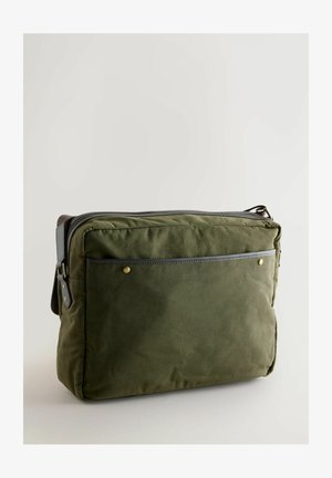 Zelená plátěná taška se zipovým uzávěrem, koženými detaily, přední kapsou a zlatými nýty. Má obdélníkový tvar a měkkou texturu.