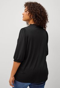 Zwarte blouse met halve mouwen, geplooide schouders en een zachte textuur. Voorzien van een rechte achterkant en een lichte glans. Gecombineerd met blauwe jeans.