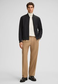 Zwarte bomberjack, witte turtleneck, beige op maat gemaakte broek, stevige zwarte schoenen; gladde stof, aansluitend ontwerp, neutraal kleurenpalet.