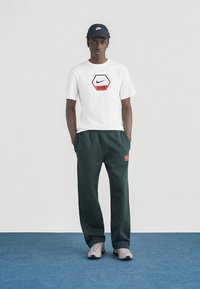 Άνδρας που φοράει λευκό μπλουζάκι Nike, σκούρο πράσινο παντελόνι φόρμας Nike, μπεζ αθλητικά παπούτσια και μαύρο καπέλο Nike, στέκεται σε μπλε πάτωμα μπροστά σε λευκό τοίχο.