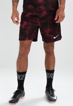 Mężczyzna ubrany w ciemnoczerwone sportowe spodenki Nike z geometrycznym wzorem, czarne skarpetki Nike oraz czarno-białe korki z różowymi kołkami, stojący na białym tle.