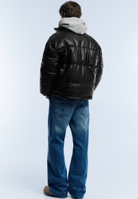 Black padded jacket with a high collar over a gray hooded sweatshirt, worn with blue jeans and brown footwear, viewed from the back.