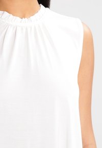 Valkoinen hihaton toppi, jossa on korkea röyhelökaulus ja pehmeä kangas, sekä löysä istuvuus, joka laskeutuu tyylikkäästi vartalolle.