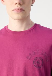 Purpurinė medvilninė marškinėliai su apvalia iškirpte ir juodu apvaliu logotipu, kuriame užrašyta "GANT 1949" krūtinės srityje.