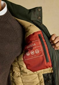 Zielona kurtka zewnętrzna z beżowym pikowanym wykończeniem; czerwona metka wewnętrzna z nazwą marki "JOULES" oraz symbolami wodoodporności. Widoczna kieszeń zapinana na zamek.